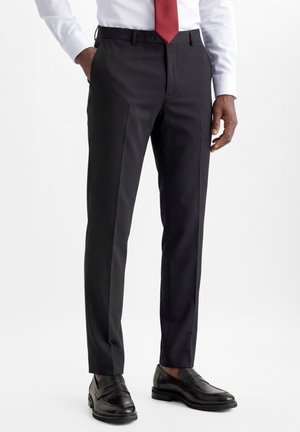 Man draagt zwarte, op maat gemaakte pantalon, zwarte leren loafers, wit overhemd en rode stropdas, staand met één hand in de zak.