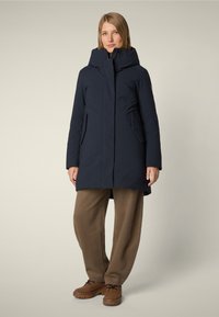 Navyblauwe, gewatteerde buitenjas met een gladde textuur, zijzakken en een middellange snit, gecombineerd met losse bruine broeken en bruine schoenen.