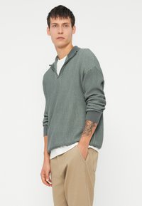 Pulover din tricot ribbed de culoare verde estompat, cu guler cu jumătate de fermoar și manșete răsfrânte, purtat peste o cămașă albă, asortat cu pantaloni bej.