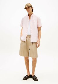 Jasnoróżowa koszula z krótkim rękawem, beżowe spodenki w paski i brązowe mokasyny. Koszula ma kołnierzyk zapinany na guziki oraz detal z logo.