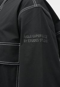 Μαύρο ύφασμα με λευκές λεπτομέρειες ραφής και κεντημένο κείμενο «AIGLE EXPERIENCE BY ÉTUDES STUDIO» στο μανίκι.