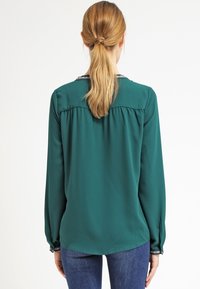 Långärmad blus i solitt grönt med rund halsringning. Har utsmyckningar på halsringningen och ärmarna, lättviktsmaterial och böjd nederkant.