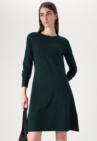 Φόρεμα μακρύ μανίκι, σκούρο πράσινο, κατασκευασμένο από μαλακό ύφασμα, με στρογγυλή λαιμόκοψη και αέρινη φούστα. Το μοντέλο κρατά μια μαύρη τσάντα.