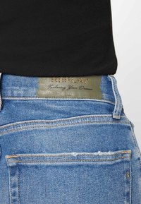 Jeans de ganga azuis com um rótulo de pele verde apresentando "REPLAY" em relevo dourado, mostrando um efeito desbotado e detalhes de costura visíveis.