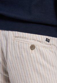 Pantaloni a righe beige con linee azzurre, dotati di chiusura a bottone e tasca posteriore. Il tessuto ha un aspetto testurizzato.