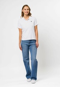 Hvit stripete polo med krage, knappdetaljer, kombinert med lyseblå flagrende jeans og hvite joggesko. Modell poserer foran.