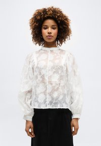 Γυναίκα με σγουρά μαλλιά που φοράει λευκή διάφανη μπλούζα με κεντήματα λουλουδιών, φουσκωτά μακριά  μανίκια και μαύρο παντελόνι, μπροστά σε απλό φόντο.