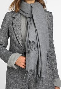 Écharpe grise en tissu doux, avec des franges aux extrémités. Portée sur un pull gris texturé et un manteau gris à motifs.
