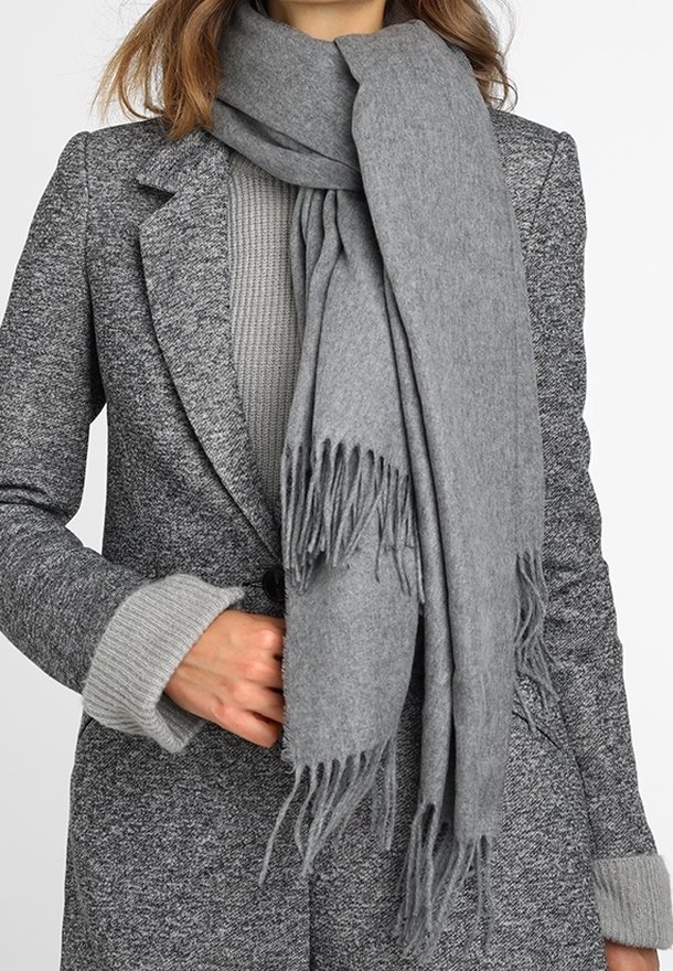 Écharpe grise en tissu doux, avec des franges aux extrémités. Portée sur un pull gris texturé et un manteau gris à motifs.