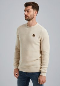 Pull en beige, texturé, avec un col rond et des manches longues. Présente un petit écusson sur la poitrine. Associé à un jean bleu foncé.