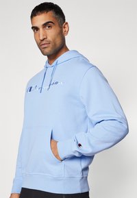 Helles blaues Baumwoll-Hoodie mit einer Fronttasche, gerippten Bündchen und einem gestickten Logo auf der Brust. Verfügt über eine Kapuze mit Zugband und eine weiche Textur.