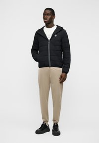 Μαύρο μαξιλαρωτό μπουφάν με φερμουάρ μπροστά, κουκούλα και λεπτομέρεια λογοτύπου, συνδυασμένο με μπεζ παντελόνια και μαύρα οξφορντ με κορδόνια. Ομαλή υφή.