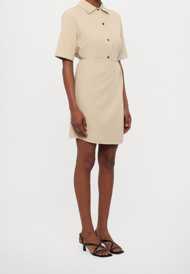 Femme portant une robe beige à manches courtes avec col, des boutons noirs et des sandales noires à brides et talons bas, debout sur un fond blanc.