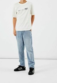 Persona vestida con una camiseta blanca de manga corta con texto, jeans de mezclilla azul claro y zapatillas negras Adidas, de pie frente a un fondo blanco liso.