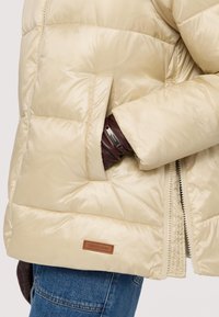 Bežová péřová bunda s prošívanou texturou, se zapínáním na zip a boční kapsou. Uvnitř se nachází hnědá nášivka s logem a viditelný rukáv pod rukávem.