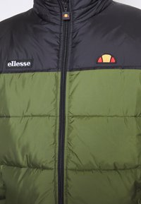 Veste matelassée noire et vert olive avec fermeture éclair centrale, logo texte « ellesse » sur le côté gauche de la poitrine, et emblème semi-circulaire rouge et jaune sur le côté droit de la poitrine.