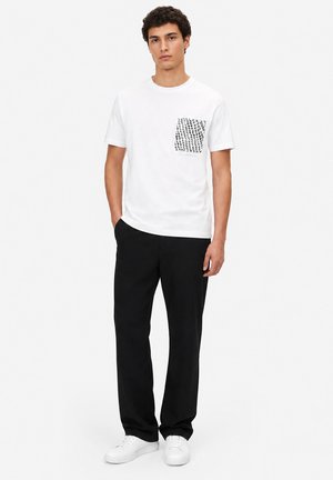 Jeune homme debout les mains dans les poches, portant un t-shirt blanc avec une poche poitrine noire à motif, pantalon noir et baskets blanches.