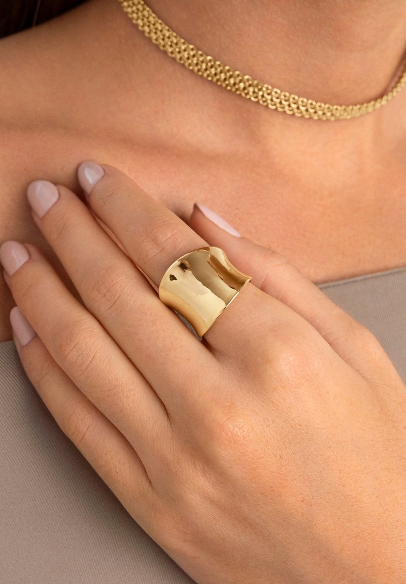 Main avec ongles rose clair portant une large bague en or brillante au doigt du milieu, reposant près de la clavicule avec un collier chaîne en or visible.