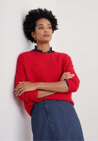 Pull en tricot rouge à rayures avec un col rond, superposé sur une blouse marine à pois colorés. Modèle portant une jupe en denim.