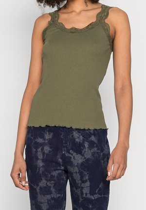 Femme portant un débardeur côtelé vert olive avec une bordure en dentelle et un pantalon bleu foncé à motif tie-dye, debout devant un fond clair uni.