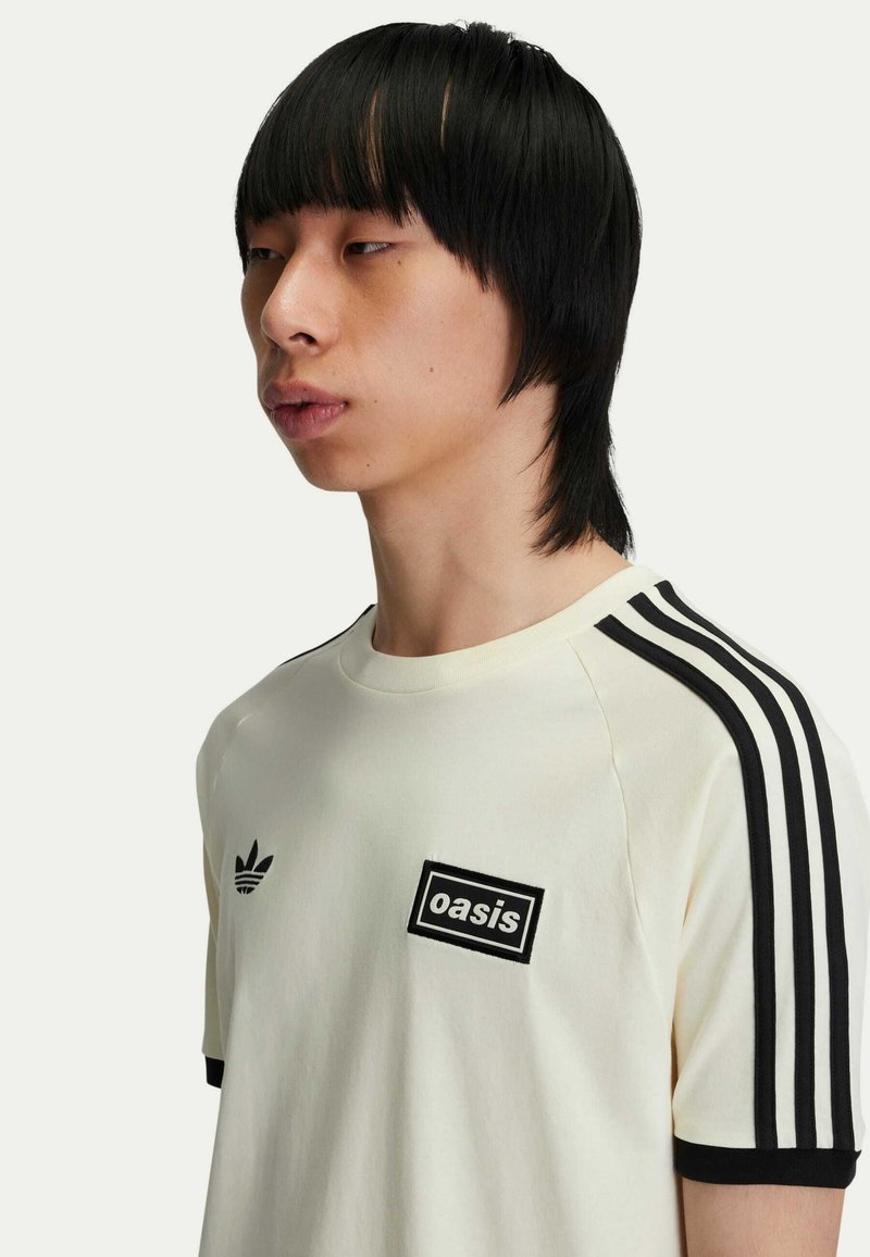 T-shirt a maniche corte color crema con dettagli a righe nere e una patch con il logo "oasis" sul petto. Materiale in cotone morbido.