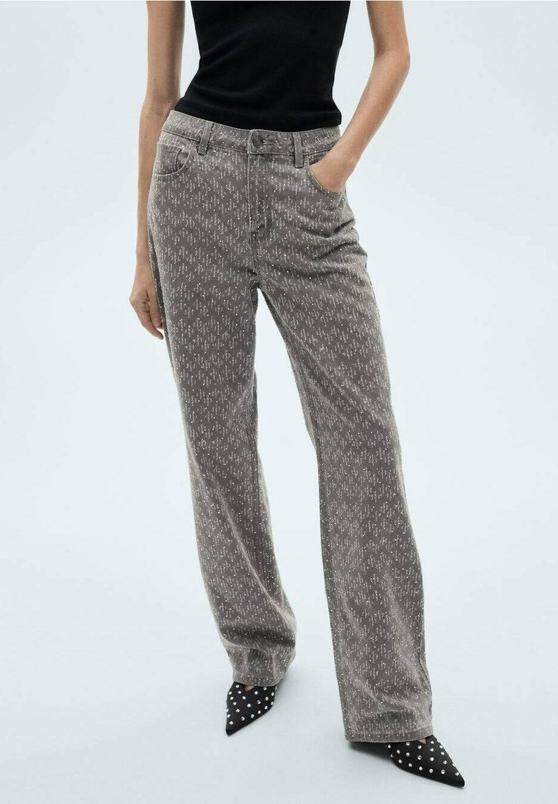 Femme portant un pantalon large à la taille haute gris à motif, un top noir sans manches, et des chaussures noires pointues à pois blancs, debout les mains dans les poches.