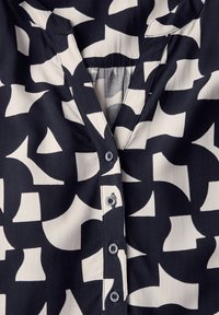 Blusa navy e bianca con motivi, scollatura a V, chiusura a bottone e forme astratte. Il tessuto appare liscio e leggero.