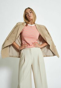 Blazer beige doppiopetto su una maglietta a righe rosse e bianche a coste, abbinato a pantaloni larghi color crema. Il look combina stili sofisticati e casual.