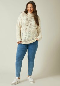 Maglione lavorato a maglia color crema con colletto alto e motivo a trecce, abbinato a jeans skinny blu e sneakers bianche con dettagli beige.