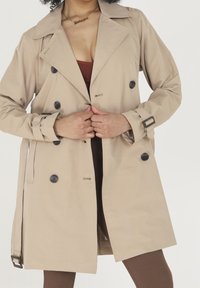 Beżowy płaszcz trenczowy z czarnymi guzikami, dwurzędowy fason, szerokie klapy i regulowany pasek. Gładki materiał z dopasowanym krojem.