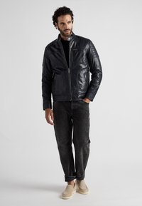 Mörkblå läderjacka med hög krage, dragkedjestängning, vadderade axelpartier och sidofickor, kombinerad med urblekta svarta jeans och slip-on skor.