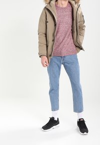 Giacca imbottita beige con bordo in pelliccia, maglione rosso screziato, jeans blu chiaro a vita alta, e sneakers nere con suola bianca, in piedi su uno sfondo chiaro.