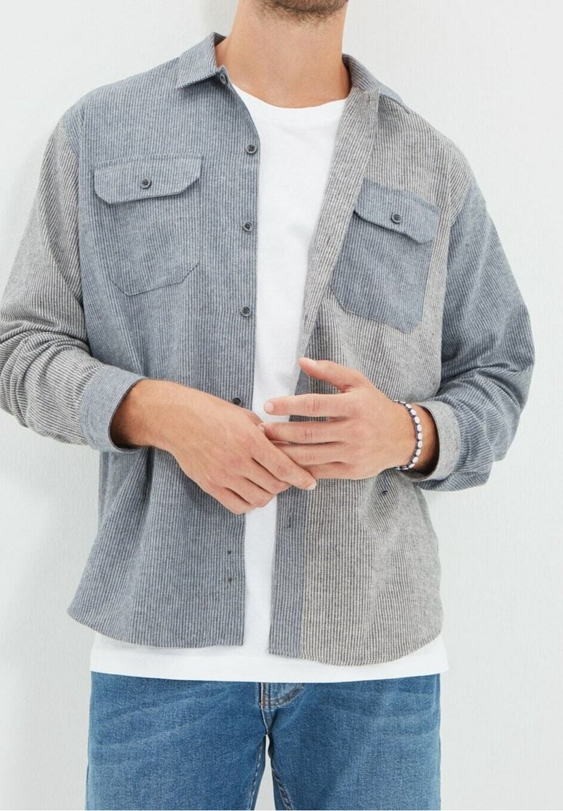 Heren overhemd met knopen in grijze ribstof, met twee borstzakken, een contrasterend paneel aan de linkerkant en een afgeronde zoom. Draagt over een wit T-shirt.