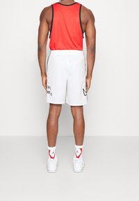 Débardeur rouge, short blanc avec des détails noirs et bleus, et baskets blanches. La vue de dos montre une musculature développée.