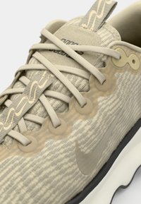Beige atletiske sko med tekstureret stofoverdel, bølget designaccenter, elastiske snørebånd, gummisål og subtilt logo nær hælen.