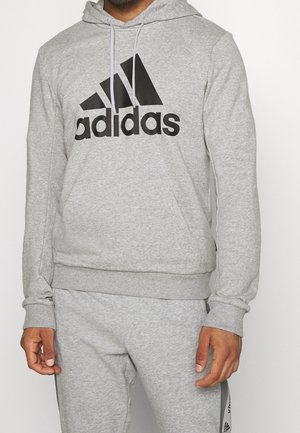 Homme portant un hoodie Adidas gris clair avec le logo noir et un pantalon de survêtement gris assorti avec une bande logo sur le côté.