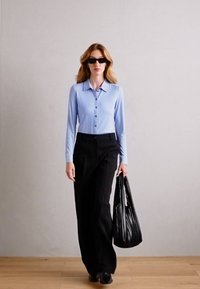 Lyseblå langermet bluse med knappestolpe, kombinert med svarte vide bukser og en teksturert svart håndveske. Enkelt og stilrent design.