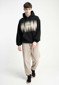 Czarna i beżowa gradientowa bluza z kapturem z napisem "INFLAMMABLE", kieszenią typu kangur oraz ściągaczami, w połączeniu z beżowymi spodniami dresowymi i czarnymi sneakersami.