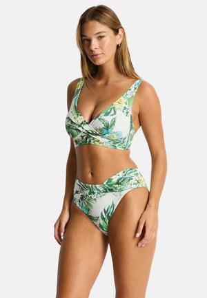 Kobieta w dwuczęściowym bikini z zielono-białym tropikalnym wzorem liści i kwiatów, stojąca na jednolitym białym tle.