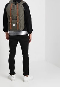 Persona con una mochila gris con correas de cuero marrón, chaqueta negra, camisa a rayas, pantalones negros y zapatillas blancas, de pie en un espacio blanco liso.