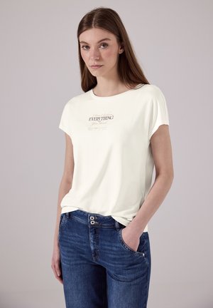 Cremefarbendes T-Shirt mit kurzen Ärmeln, mit einem vorn bedruckten Textdesign. Kombiniert mit blauen Jeans, die eine lockere Passform zeigen.