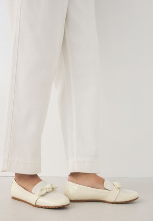 Personne portant un pantalon écru et des mocassins crème avec un détail noué à l'avant, debout sur un sol clair.