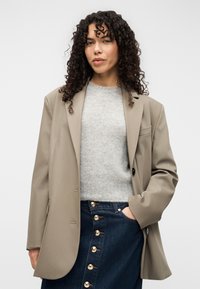 Μπεζ oversized σακάκι με ένα κουμπί, φορεμένο πάνω από ανοιχτό γκρι πουλόβερ. Συνδυάζεται με τζιν φούστα με χρυσά κουμπιά και μπροστινή τσέπη.