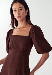 Rjava obleka z napihnjenimi rokavi z kvadratnim izrezom, nagubano na ramenih in s prilegajočim pasom, izdelana iz lahkotnega blaga.