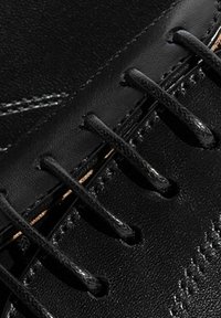 Primo piano di una scarpa in pelle nera, con lacci prominenti, superficie texture e cuciture bianche a contrasto lungo i bordi.