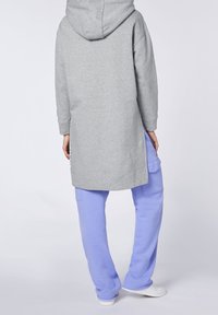 Felpa grigia con cappuccio e orlo lungo, abbinata a pantaloni della tuta viola chiaro con tasche laterali e polsini elasticizzati. Vestibilità rilassata e tessuto morbido.
