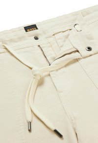 Krémové kalhoty s částečně rozepnutým zipem, kovovým knoflíkem a okem, a s uzlovou šňůrkou v pase.