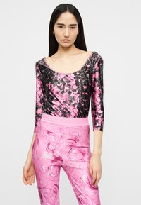 Γυναίκα με μακριά σκούρα μαλλιά, που φοράει μπλούζα με μοτίβο από ροζ και μαύρες λέπια και ροζ ψηλόμεσο παντελόνι με μπαρόκ σχέδια.