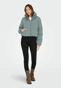 Korte, pufferjack in gedempt groen corduroy met een rits aan de voorkant, capuchon en zijzak, gecombineerd met zwarte skinny jeans en bruine enkelboots.