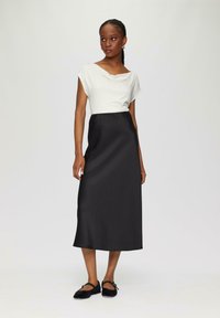 Schwarzes Satin-Maxi-Kleid mit einem weißen drapierten Oberteil, das einen gewellten Taillenbund hat, und kombiniert mit schwarzen flachen Schuhen.